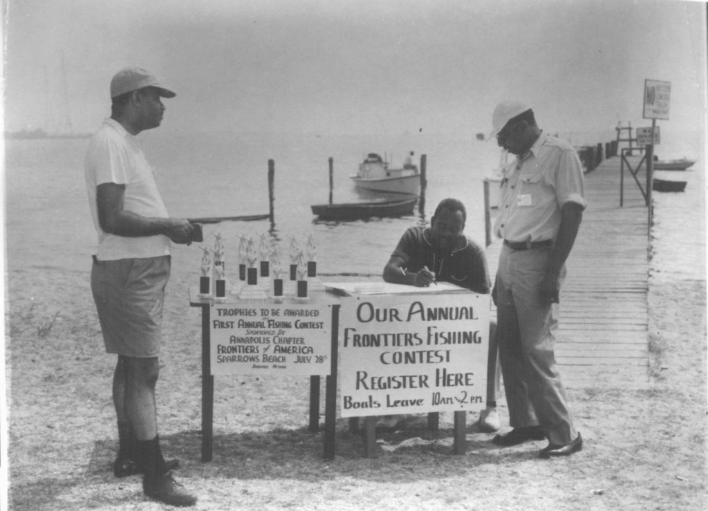 three men next to tables with fishing contest sign and trophies